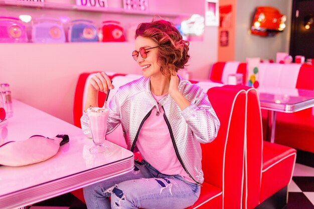 Mujer en retro vintage american 50's cafe sentado en la mesa bebiendo un cóctel de batido de leche en estilo hipster con gafas de sol rosas y traje de chaqueta plateada divirtiéndose riendo