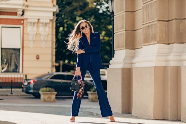 Mujer rica de lujo vestida con elegante traje azul elegante caminando en la ciudad en un día soleado de otoño sosteniendo el bolso