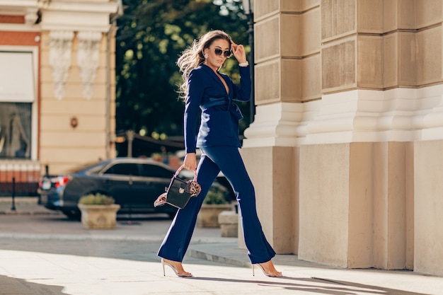 Mujer rica de lujo vestida con elegante traje azul elegante caminando en la ciudad en un día soleado de otoño sosteniendo el bolso