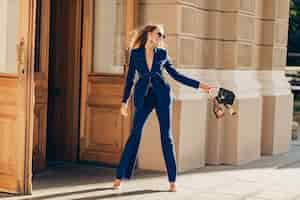 Foto gratuita mujer rica de lujo vestida con elegante traje azul elegante caminando en la ciudad en un día soleado de verano con bolso