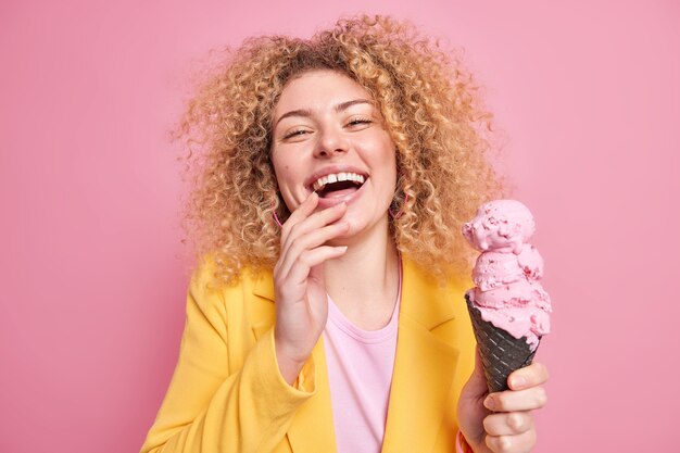 La mujer se ríe alegremente sostiene un helado de cono de sabor a fresa en gofre negro tiene un estado de ánimo optimista viste una elegante chaqueta amarilla