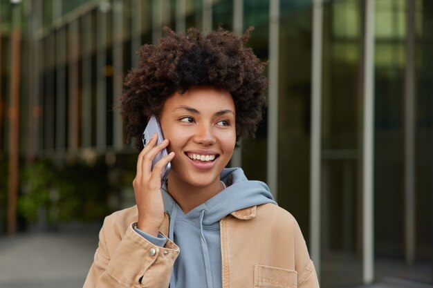 mujer se ríe mientras llama a teléfonos inteligentes conversaciones en itinerancia tiene el pelo rizado vestido con ropa casual posa afuera hace comunicación internacional