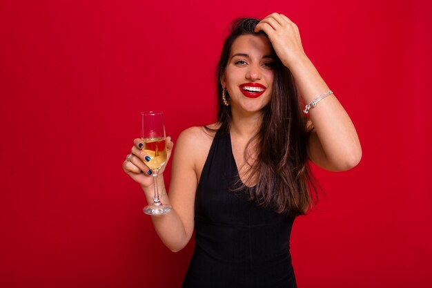 Mujer riendo con adorable sonrisa vistiendo ropa negra con lápiz labial rojo sosteniendo una copa de champán posando sobre pared roja