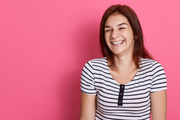 Mujer riendo con cabello oscuro posando aislada sobre pared rosada, niña feliz con camiseta a rayas, expresando felicidad y alegría. Copie el espacio para publicidad.