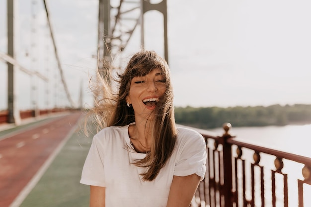 Mujer riendo emocionada con una sonrisa maravillosa con camiseta blanca si los ojos cerrados y sonriendo y disfrutando de la mañana de verano caminando por el puente en la ciudad