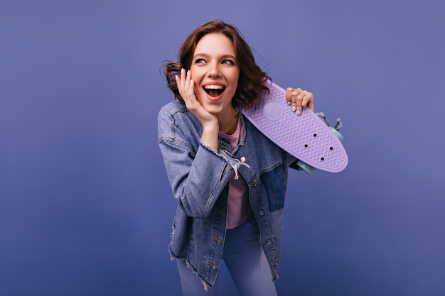 Mujer riendo feliz con peinado ondulado divirtiéndose. Filmación en interiores de encantadora chica rizada en chaqueta de mezclilla con longboard.