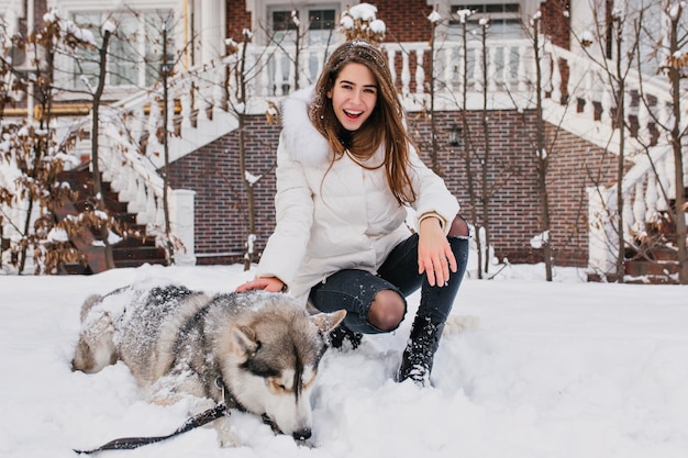 Mujer riendo feliz con el pelo lacio sentado en la nieve junto a su perro. mujer guapa en jeans y chaqueta blanca posando con husky después de caminar en la mañana de invierno.