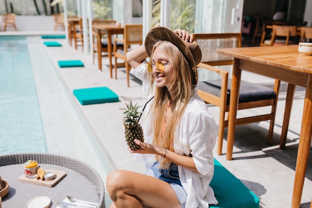 Mujer riendo con gafas de sol amarillas bebiendo cóctel de frutas y tomando el sol.