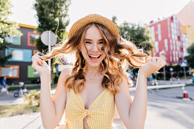 Mujer riendo glamorosa jugando con su cabello rubio ondulado. Foto al aire libre de niña adorable en vestido amarillo y sombrero vintage.