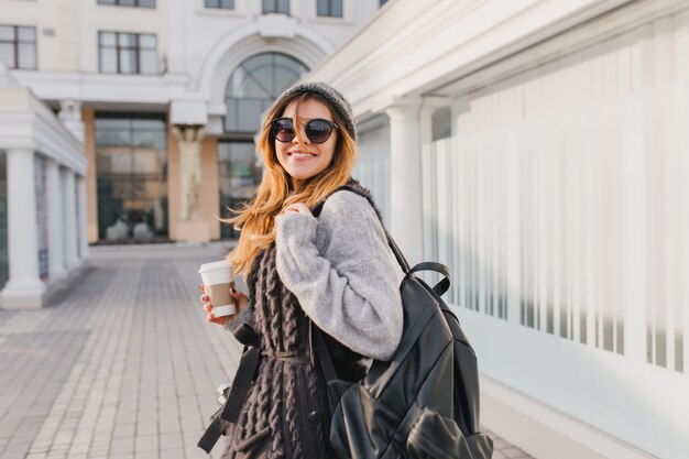 Mujer riendo con mochila negra caminando por la ciudad y tomando café en buen día. Retrato al aire libre del viajero femenino sonriente en suéter y sombrero posando