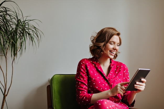 Mujer riendo sentada en un sillón y usando tableta digital. Filmación en interiores de una mujer bonita en pijama con gadget y sonriendo.