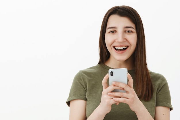 Mujer riendo sobre una divertida foto tuya tomada a través de un teléfono inteligente sosteniendo el teléfono móvil en las manos sonriendo ampliamente divertido y despreocupado divirtiéndose mientras posa contra la pared blanca con el dispositivo