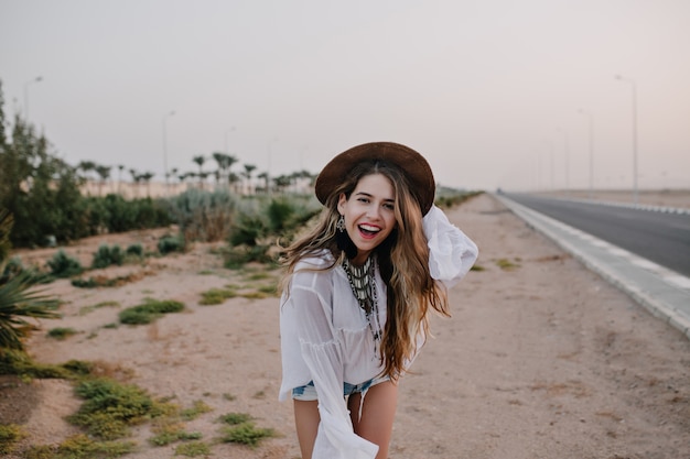 Mujer riendo con sombrero marrón de moda y blusa blanca divirtiéndose de pie en la arena junto a la carretera. Retrato de encantadora joven de pelo largo bailando afuera y posando con una sonrisa, disfruta de las vacaciones