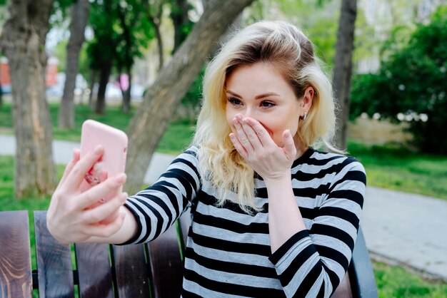 Mujer riendo tomando selfie con smartphone mientras está sentado al aire libre en un banco en la parte superior a rayas