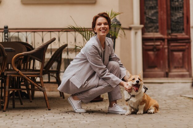 Mujer riendo en traje gris riendo y jugando con perro. Encantadora dama de pelo corto en elegante chaqueta y pantalón sonriendo y posando con corgi