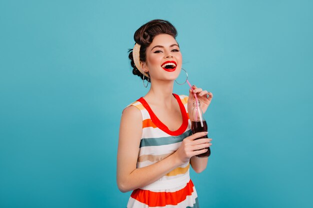 Mujer riendo en vestido rayado bebiendo refrescos. Disparo de estudio de feliz chica pinup con bebida aislada sobre fondo azul.