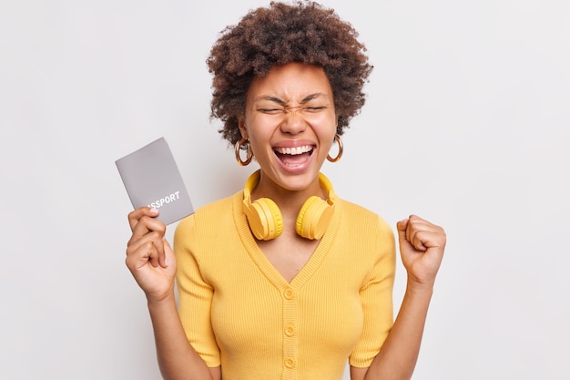 Foto gratuita la mujer rizada alegre aprieta el puño se regocija en el futuro viaje al extranjero durante las vacaciones sostiene el pasaporte lleva auriculares alrededor del cuello vestida con ropa amarilla aislada sobre una pared blanca