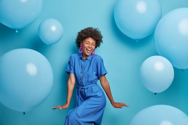 La mujer rizada alegre y despreocupada baila feliz, vestida con un vestido azul, se enfría en la fiesta alrededor de globos de helio inflados, se siente juguetona, disfruta de sus vacaciones favoritas, tiene un ambiente festivo optimista. Momento de alegria