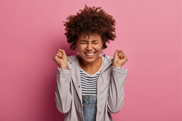 Foto gratuita la mujer rizada, alegre, emotiva y étnica, aprieta los puños, celebra la victoria, sonríe ampliamente, está muy animada, cierra los ojos, usa un anorak informal, se regocija por lograr el objetivo, posa contra la pared rosa.