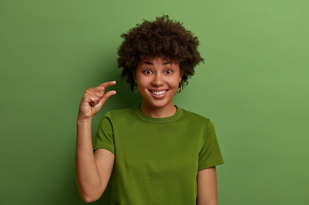 La mujer rizada, alegre y positiva, de piel oscura, da forma a algo muy pequeño con los dedos, demuestra una pequeña disminución en el precio o el salario, hace gestos que no son un objeto grande, sonríe con los dientes, prevalece el color verde