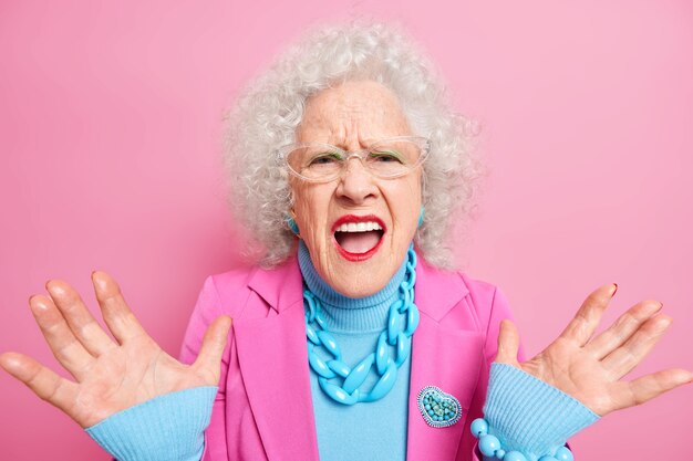Mujer rizada anciana decepcionada levanta palmas exclama en voz alta expresa emociones negativas viste ropa elegante lápiz labial rojo y maquillaje