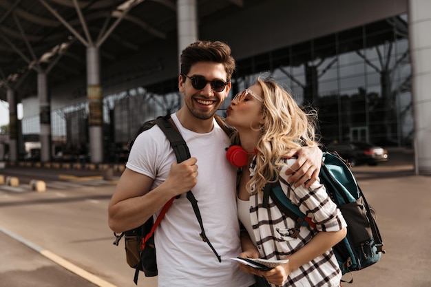 Mujer rizada besa a su novio cerca del aeropuerto Chica atractiva con camisa a cuadros y hombre moreno con camiseta blanca posa de buen humor y sostiene mochilas