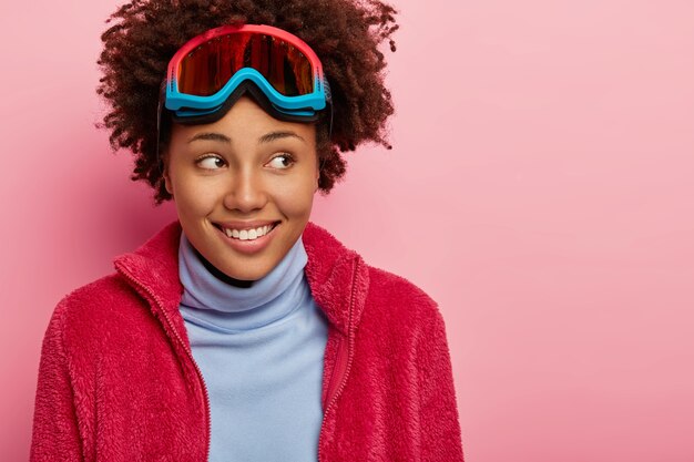 Mujer rizada complacida vestida con ropa de invierno, lleva gafas de esquí en la frente, mira con alegría a un lado, modelos sobre una pared rosa.