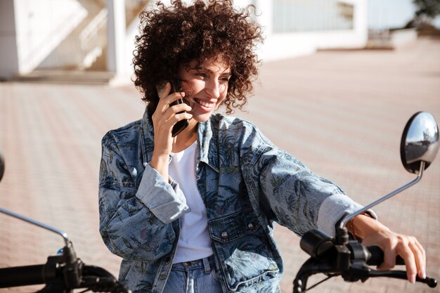 Mujer rizada contenta sentada en moto moderna al aire libre y hablando por el teléfono inteligente