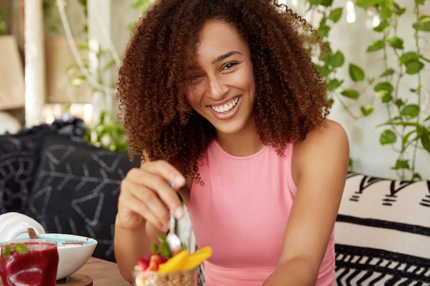 Mujer rizada con expresión alegre, come delicioso postre, está de buen humor, pasa tiempo libre en una acogedora cafetería, disfruta de una sabrosa ensalada de frutas. Atractiva hembra descansa después de una excursión sola