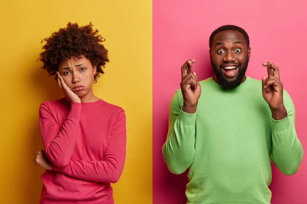 La mujer rizada de fatiga molesta se ve triste, su novio está feliz cerca, mantiene los dedos cruzados, cree en la buena fortuna, usa un jersey verde, se para contra la pared amarilla y rosa