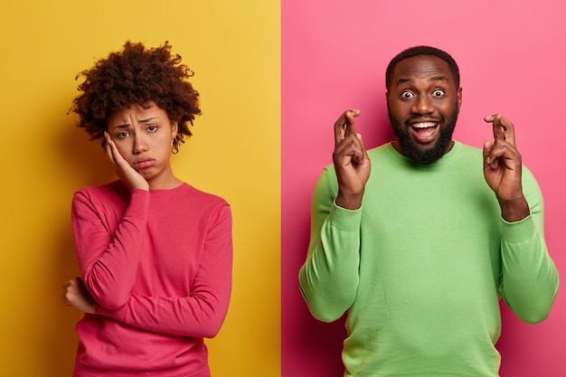 Foto gratuita la mujer rizada de fatiga molesta se ve triste, su novio está feliz cerca, mantiene los dedos cruzados, cree en la buena fortuna, usa un jersey verde, se para contra la pared amarilla y rosa