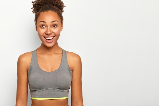 La mujer rizada feliz tiene una sonrisa brillante, muestra los dientes blancos, está vestida con un sostén deportivo informal, hace ejercicio o hace ejercicio durante el tiempo libre, modelos de interior