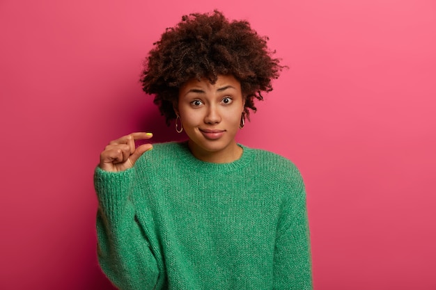 La mujer rizada mide algo diminuto, dice que tiene pequeños problemas, da forma a objetos pequeños, viste un suéter verde, no parece impresionada, posa sobre una pared rosada. Concepto de tamaño y lenguaje corporal