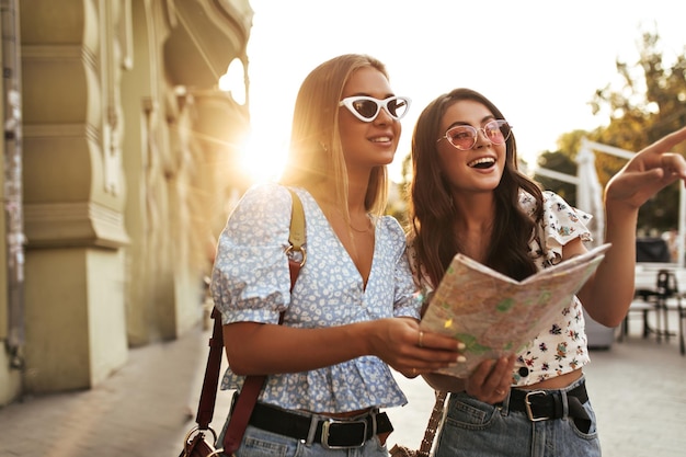 Mujer rizada morena bastante bronceada y encantadora chica rubia con blusas florales y gafas de sol sostienen un mapa y caminan en la hermosa ciudad