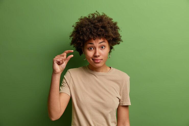 Foto gratuito mujer rizada no impresionada muestra algún gesto más o poco, demuestra pocas pulgadas o centímetro, pequeña cantidad o signo de baja escala, dresseed en ropa casual, aislado en la pared verde