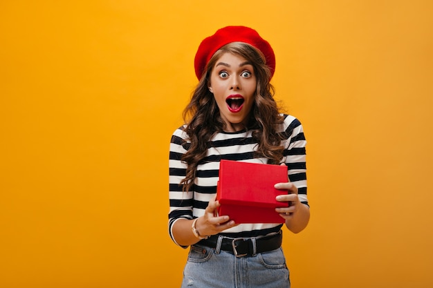 La mujer rizada parece sorprendida después de abrir la caja de regalo. Chica sorprendida en boina roja y blusa a rayas mirando a cámara se regocija presente.