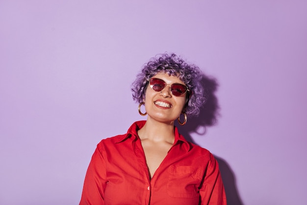 Mujer rizada de pelo corto con elegantes gafas brillantes, camisa roja con manga larga y sonrisas en lila aislado.