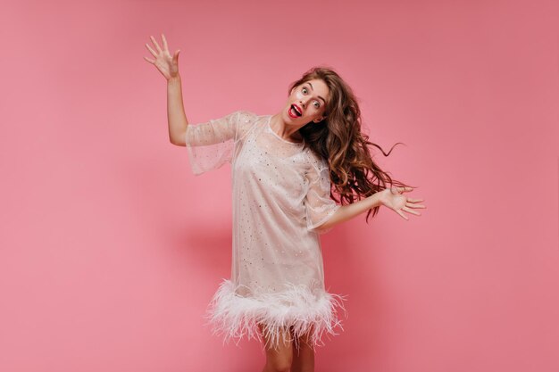 Mujer rizada de pelo largo con vestido blanco sonríe y baila sobre fondo rosa Chica genial con ropa elegante se mueve aislada