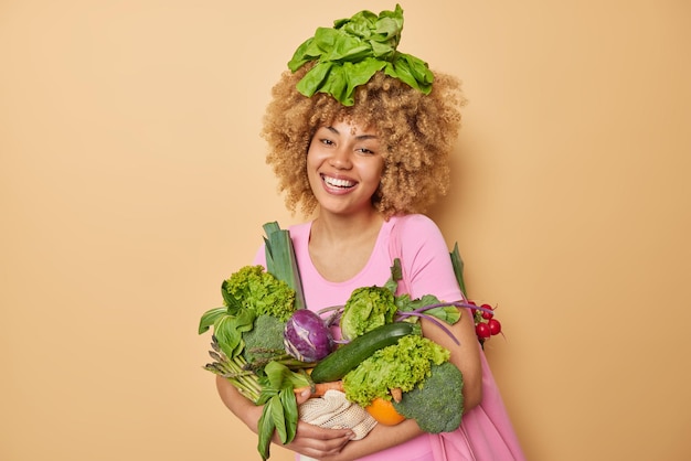 Una mujer rizada positiva posa con vegetales verdes frescos llenos de vitaminas contenta de tener sonrisas ricas en la cosecha con gusto usa una camiseta casual aislada sobre fondo marrón Concepto de alimentación saludable