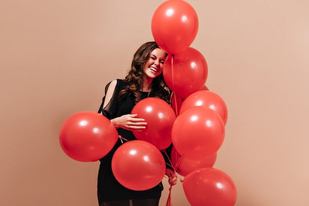 Foto gratuita mujer rizada positiva en traje negro se ríe y posa con globos rojos sobre fondo aislado.