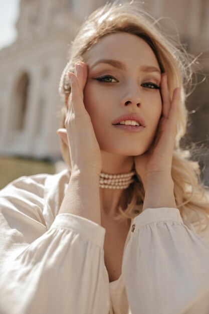 Mujer rizada rubia de ojos grises con blusa blanca y collar de perlas toca la cara, mira a la cámara y camina al aire libre