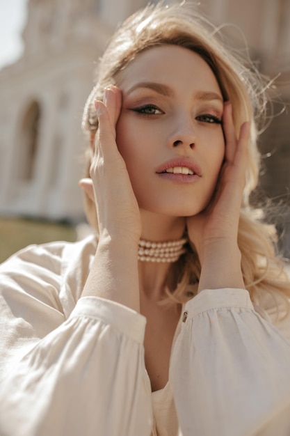 Mujer rizada rubia de ojos grises con blusa blanca y collar de perlas toca la cara, mira a la cámara y camina al aire libre