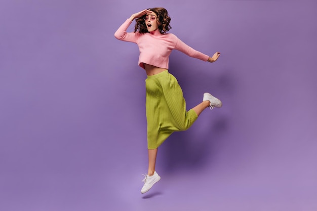 Mujer rizada saltando sobre fondo púrpura Joven chica alegre en falda verde y suéter rosa mira hacia otro lado aislado