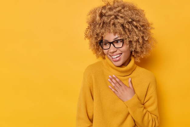 La mujer rizada sincera positiva mantiene la mano en el pecho mira hacia otro lado alegremente sonríe ampliamente se ríe de algo divertido lleva gafas y un jersey casual aislado sobre fondo amarillo. Concepto de emociones