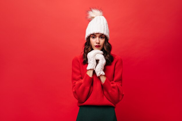 Mujer rizada en suéter rojo, gorro de punto y guantes mira al frente