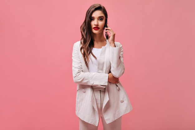 Mujer rizada en traje blanco habla por teléfono. Señora elegante con labios rojos en traje de moda ligero sobre fondo rosa aislado.