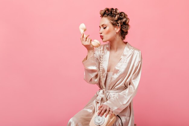 Mujer rizada en traje de casa de seda se sienta en una silla en la pared rosa y besa el teléfono
