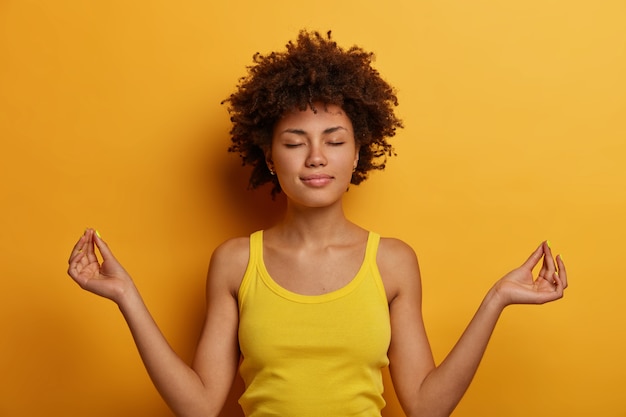 La mujer rizada tranquila y pacífica se encuentra en la postura del loto, alcanza el nirvana, practica yoga o meditación, mantiene los ojos cerrados, se viste con ropa casual, se aísla sobre una pared amarilla, hace un signo bien o zen