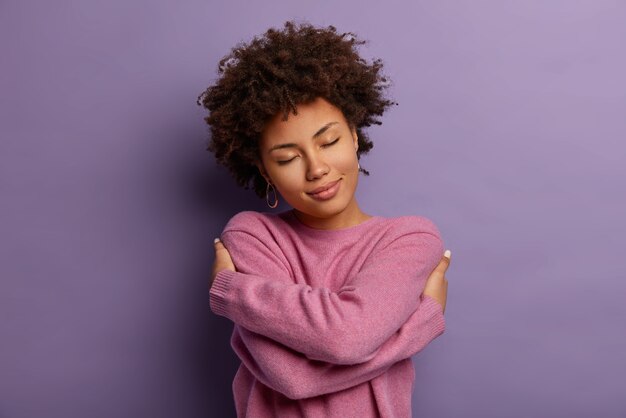 La mujer romántica se abraza a sí misma, mantiene los brazos cruzados sobre el cuerpo, toca los hombros, disfruta de la comodidad, usa un suéter, inclina la cabeza, tiene una alta autoestima, se ama a sí misma, aislada en la pared púrpura Foto gratis