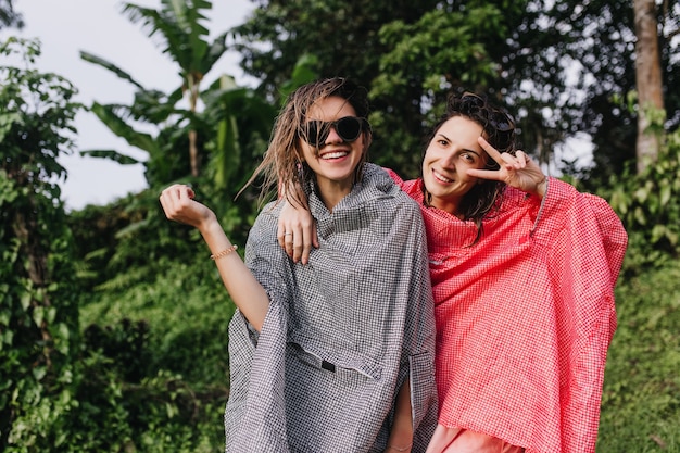 Mujer romántica en gabardina rosa posando con el signo de la paz estando cerca de un amigo. Tiro al aire libre de mujeres inspiradas descansando en el bosque.